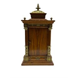 Late 19th century American mantle clock in a light oak case, raised pediment with a recessed balustrade frieze, pierced brass crown and finial, with arched rectangular conforming panels to the sides of the case, four wooden columns with capitals to each corner on a stepped plinth, circular repoussé brass dial with applied blue on white porcelain Roman numerals and Gothic steel hands, brass bezel with an egg and dart slip and flat bevelled glass, spring driven Ansonia eight-day striking movement with a recoil anchor escapement striking the hours and half-hours on a coiled gong, with fine pendulum regulation. With key and pendulum.

