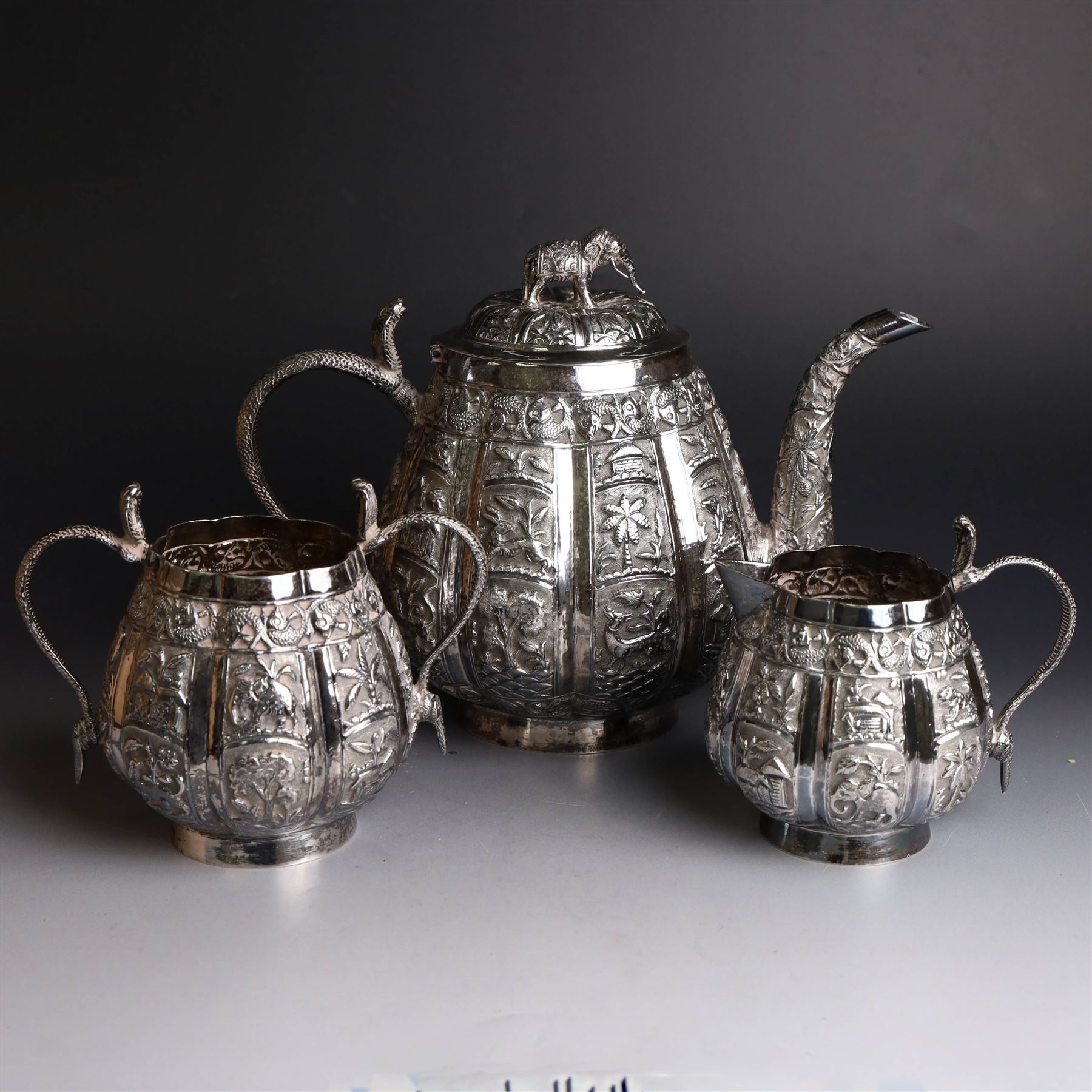Three piece Indian silver tea service, comprising teapot, twin handled open sucrier and cream jug, each with embossed animal decoration with fish border, serpent handles and elephant finial, teapot H17cm