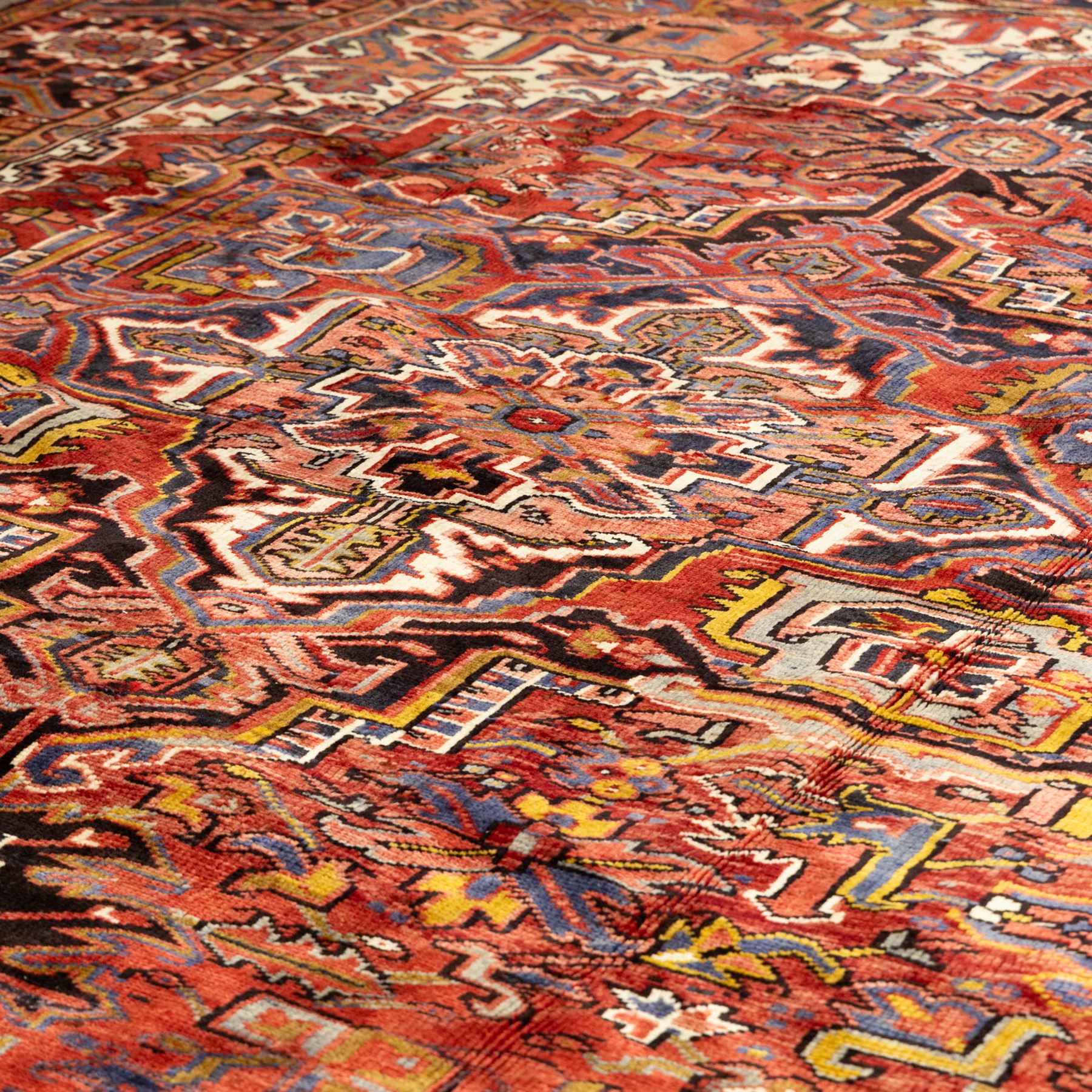 North West Persian Heriz hand-knotted carpet, madder red ground, with a large central geometric medallion with pendants, surrounded by angular foliate and floral motifs incorporating stylised rosettes, palmettes and serrated vine work, enclosed by ivory spandrels and a dark navy border with repeating geometric flowerheads and angular meandering vines, flanked by narrow patterned guard stripes, with cotton fringes