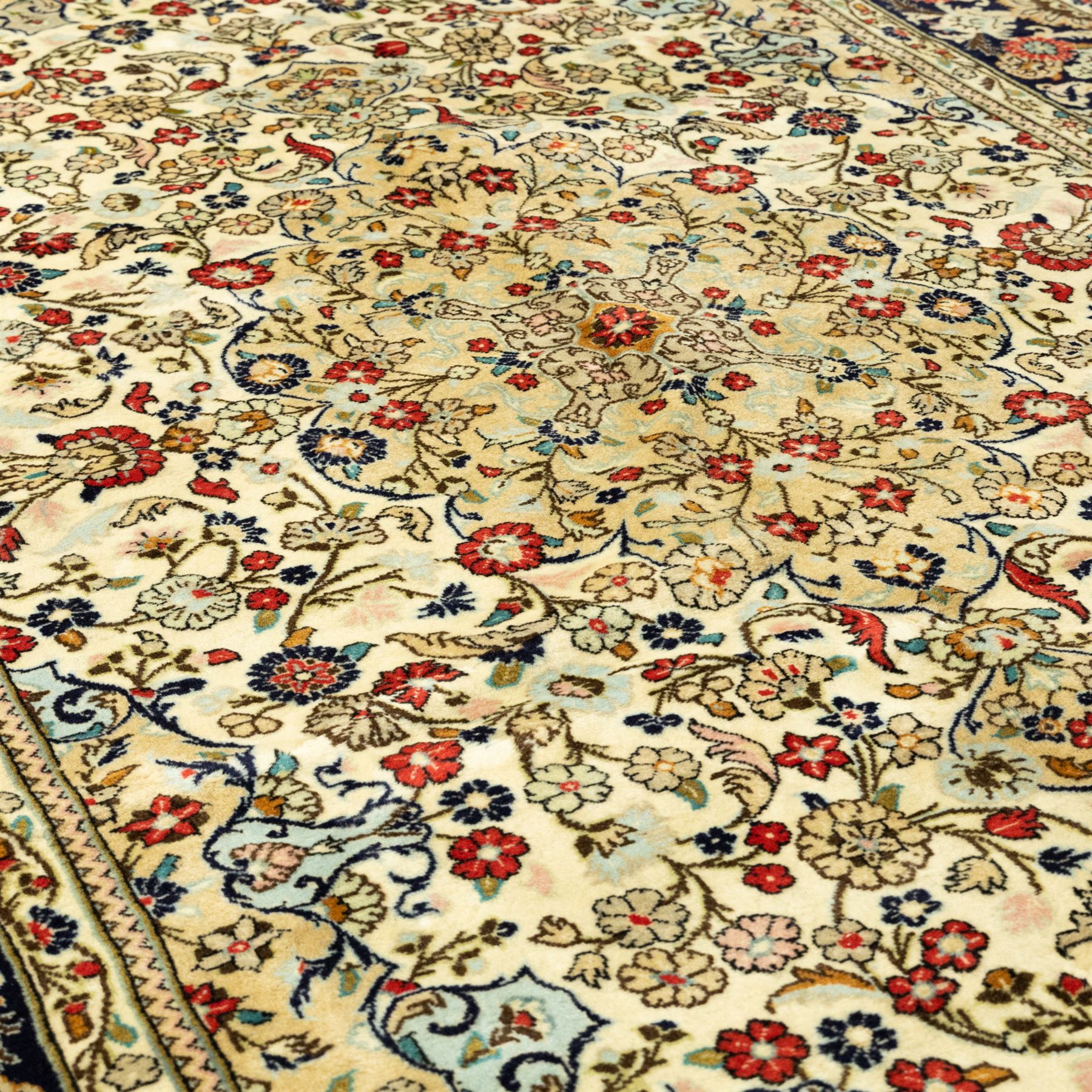 Central Persian Kashan hand-knotted carpet, ivory ground, with an all-over floral field centred by a subtle shaped medallion, surrounded by scrolling foliate and floral motifs incorporating rosettes, palmettes and delicate vine work, enclosed by a dark navy border with repeating palmettes and meandering vines, flanked by floral guard stripes, with cotton fringes