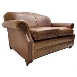 Traditional shape two-seat sofa, shaped back and rolled arms upholstered in tan leather with stud work bands, on compressed bun feet with castors 