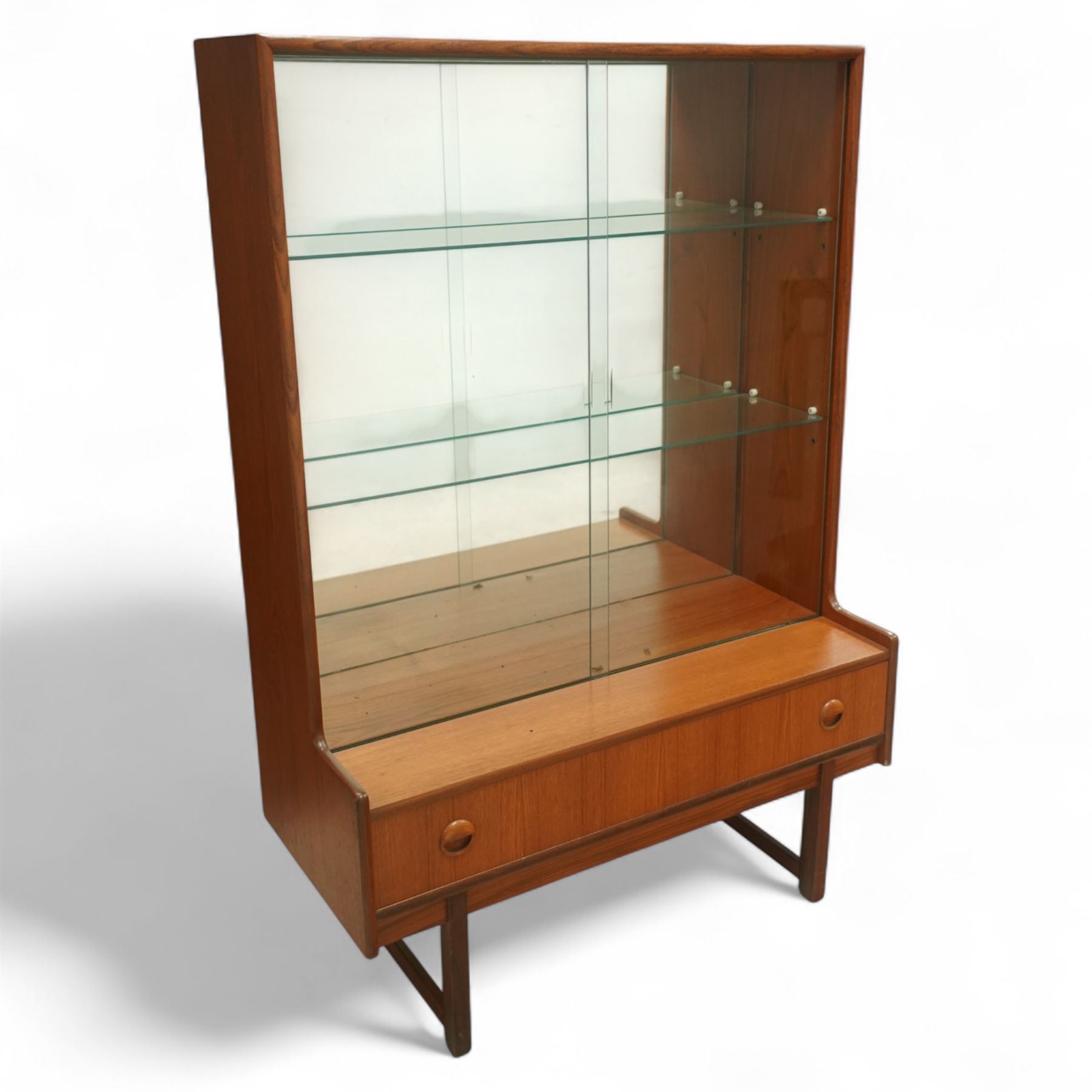 Turnidge of London mid-century teak display cabinet, rectangular top with a pair of glass sliding doors enclosing two adjustable shelves with mirrored back, projecting lower section incorporating one full-width drawer with recessed circular handles, raised on square-section legs with lateral stretchers  