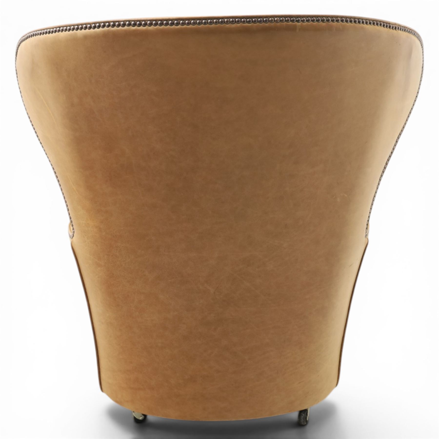 20th century tan leather armchair, the curved wing back above low square arms, shaped loose seat cushion with piped edge, close-nailed side detail, raised on concealed castors