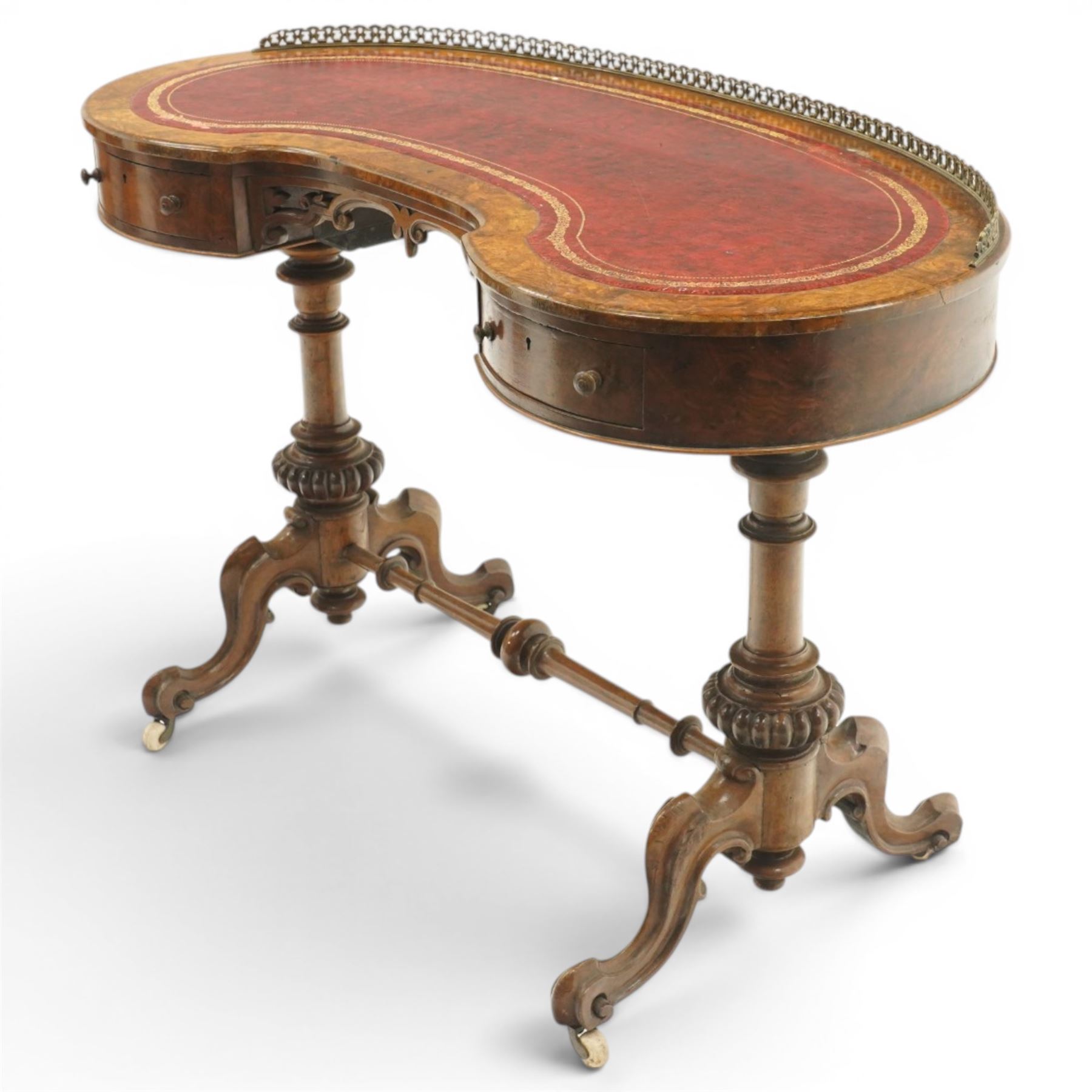 Victorian figured burr walnut kidney-shaped writing table, shaped top with pierced raised bras gallery back over inset red leather writing surface, fitted with two drawers flanking a pierced and carved scrolling apron, raised on turned and gadrooned twin pedestals with scroll-moulded cabriole supports with ceramic castors, united by ring-turned stretcher