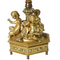 Second Empire style gilt metal table lamp, the hexagonal plinth base with applied foliate decoration, upon six turned feet, supporting three seated allegorical putti, the first modelled with a tablet and stylus representing literature, the second with hammer representing sculpture, and the third with Corinthian column capital representing Architecture, each intersected by a stylised scroll with central flower head panel, mounted upon an onyx and gilt metal Corinthian column with wrythen flower moulded and part fluted base, supporting a cream octagonal shade, overall H86cm