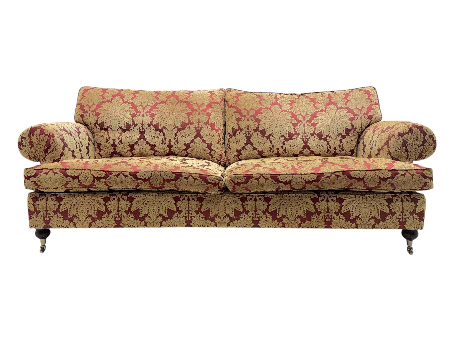 Duresta - pair of Grande three-seat sofas, traditional shaped with rolled arms and upholstered loose cushions, in red ground fabric with raised gold foliate pattern, on turned feet with brass castors 