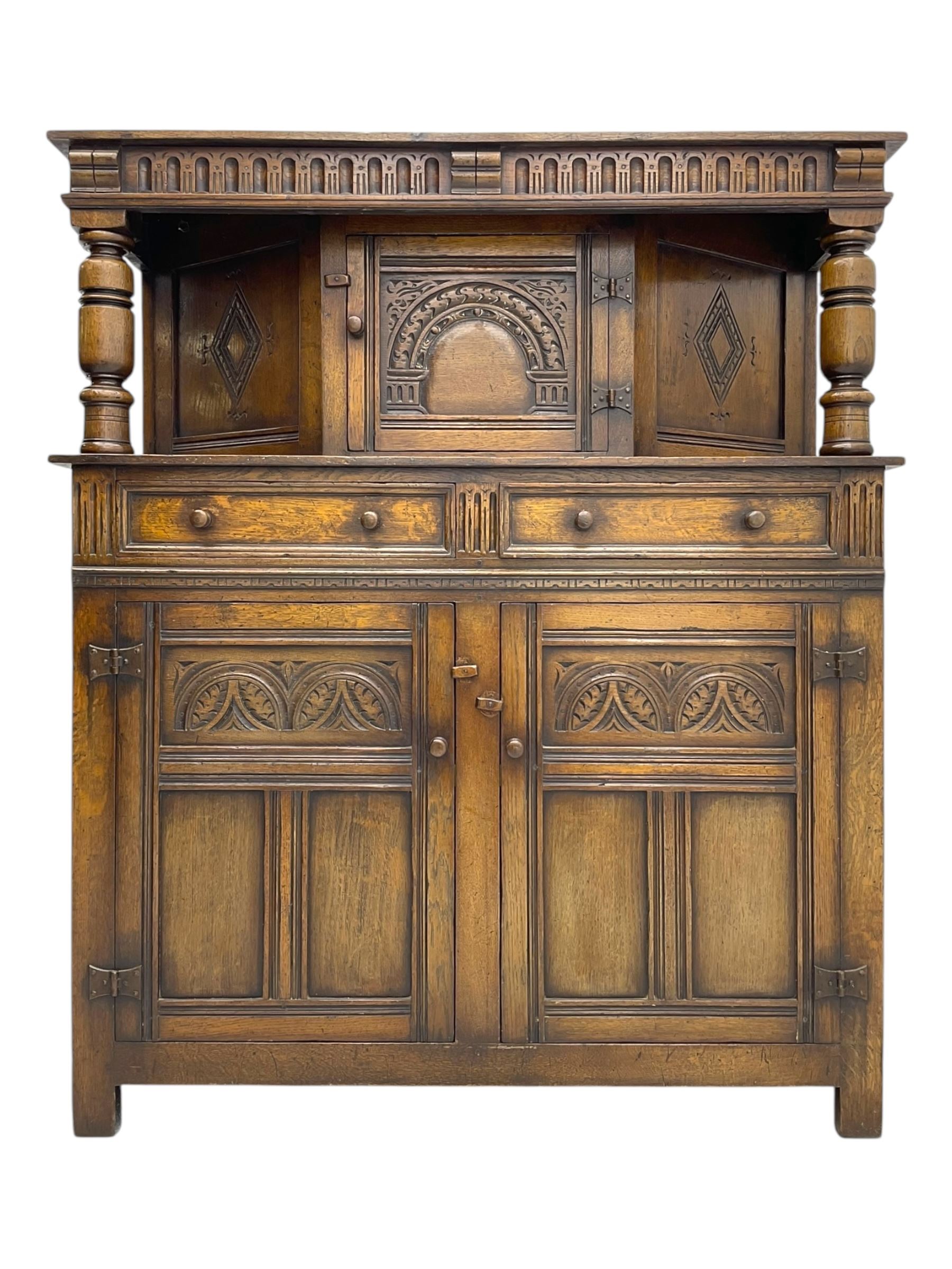 Jacobean design oak court cupboard, upper section with carved arched panelled cupboard door flanked by turned columns, beneath carved and moulded frieze, lower section fitted with two drawers over two cupboard doors with gothic arched panel decoration, raised on stile supports