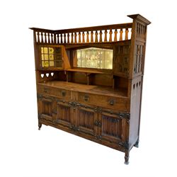 Shapland and Petter - Arts & Crafts period oak dresser, raised gallery top with spade balustrade, central bevelled mirror flanked by two cabinet, astragal glazed doors with yellow tinted glass and bullseye panes, the sides with pierced heart shaped decoration, fitted with two drawers and two double cupboards, enclosed by linenfold panelled doors,  copper work strap hinges and fixtures, on shaped spade feet, the locks stamped 'S&P'