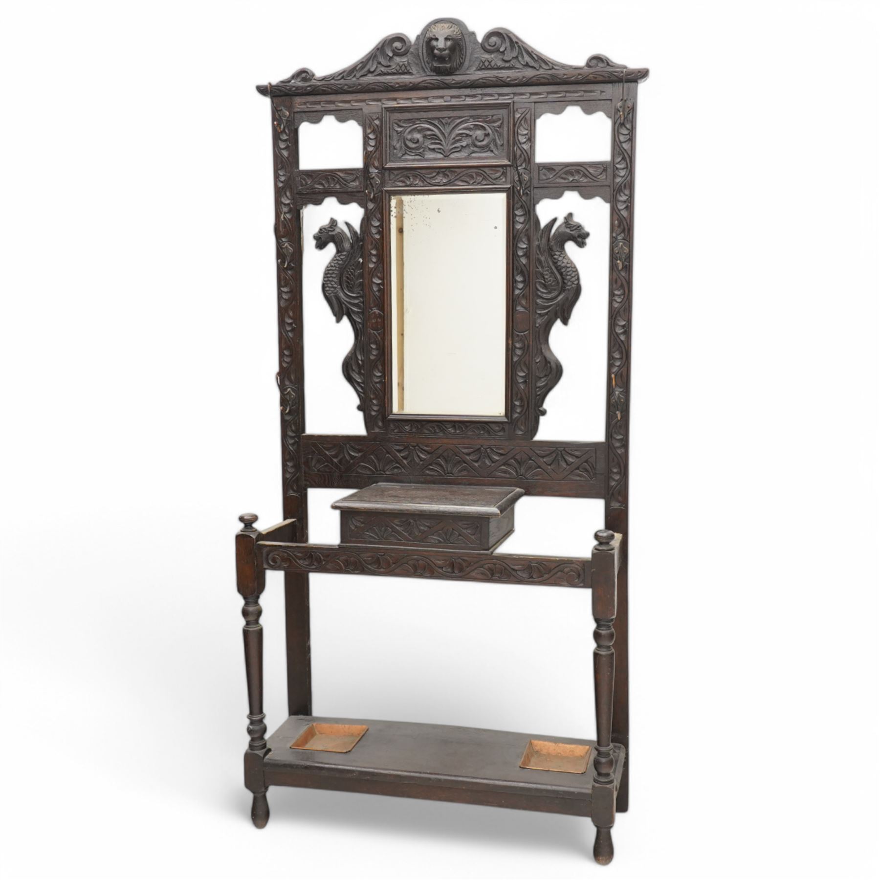 Late Victorian heavily carved oak hall stand, the scrollwork pediment with carved lion's head, bevelled mirror plate flanked by carved dragon motifs, over hinged storage compartment and umbrella stands with drip trays, on turned front supports