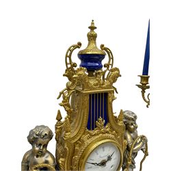 A 20th century continental gilt metal Lyre mantle clock with a pair of matching six light candelabra, spring driven movement housed in a gilt drum case on a brass and cobalt blue base with paw feet, two bronzed metal figures of mythological cherubs with animal legs and cloven hooves supporting a festooned garland swag, eight-day twin barrel striking movement with a floating lever balance escapement, striking the hours and half-hours on two bells, white enamel dial with roman numerals, minute track and pierced steel hands, dial inscribed “Imperial”. 