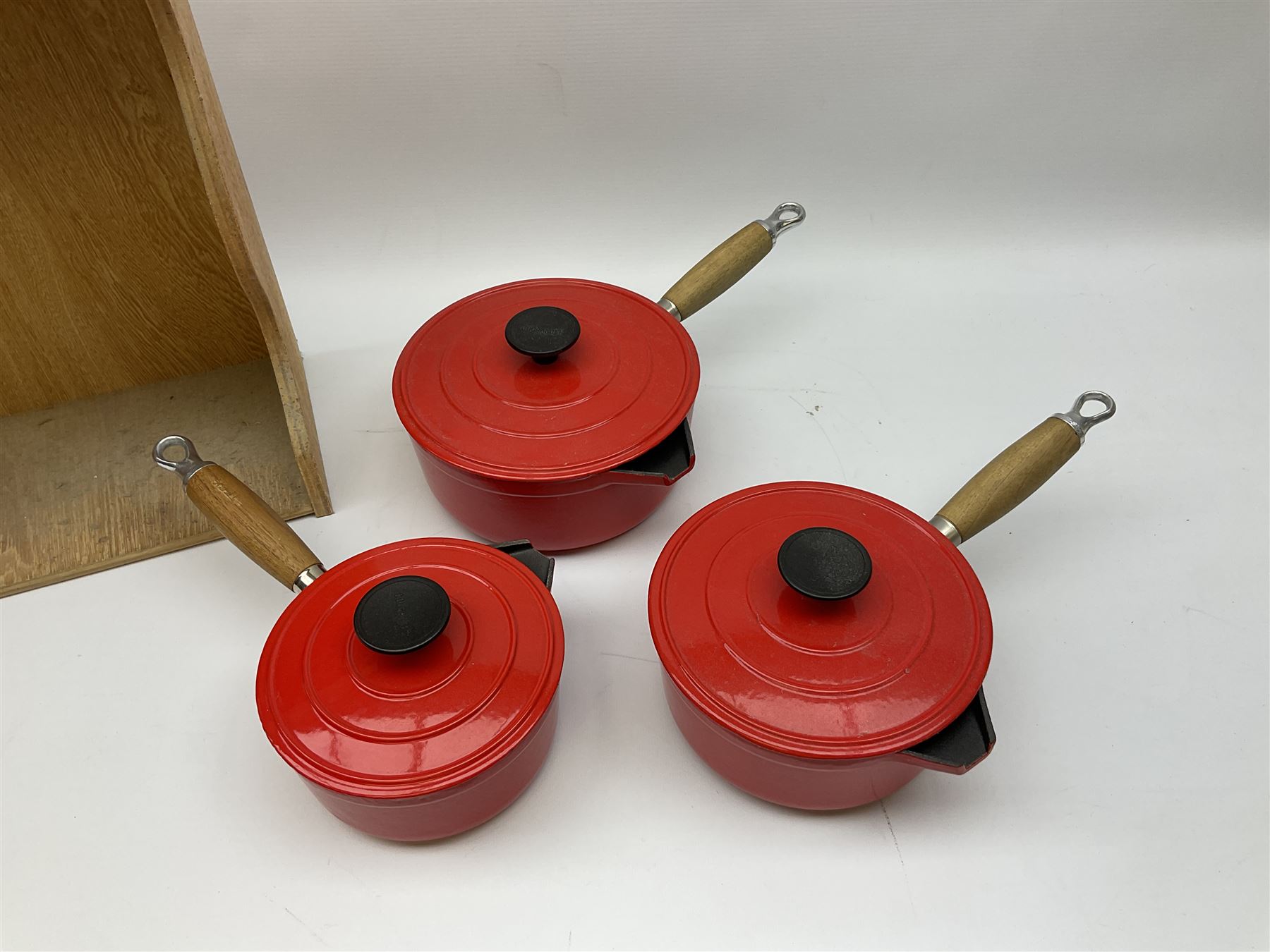 Le Creuset 'Volcanic Orange' cast iron and enamel milk pan with wood handle together with a three piece red cast iron Cousances saucepan set of graduating form, with lids housed in original wooden hanging stand and further Cousances lidded casserole dish and pan
