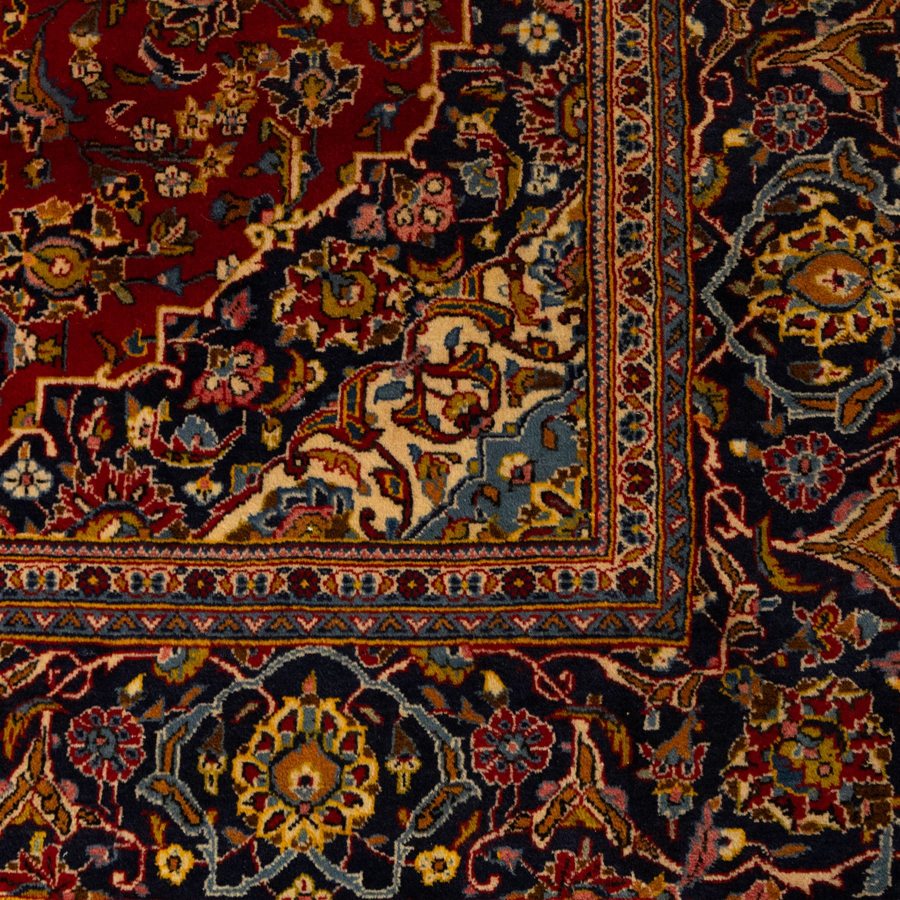 Central Persian Kashan hand-knotted carpet, crimson red field with a central shaped palmette medallion with pendants, filled with dense scrolling and floral motifs, enclosed by a navy blue border decorated with palmettes and meandering vine scrolls, flanked by multiple guard stripes, with cotton fringe ends 