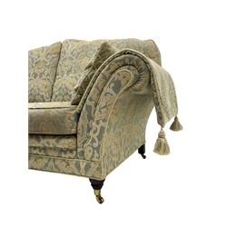 Traditional shape two seat sofa, upholstered in repeating foliate pattern fabric, with side cushions and arm covers, on turned feet with brass cups and castors