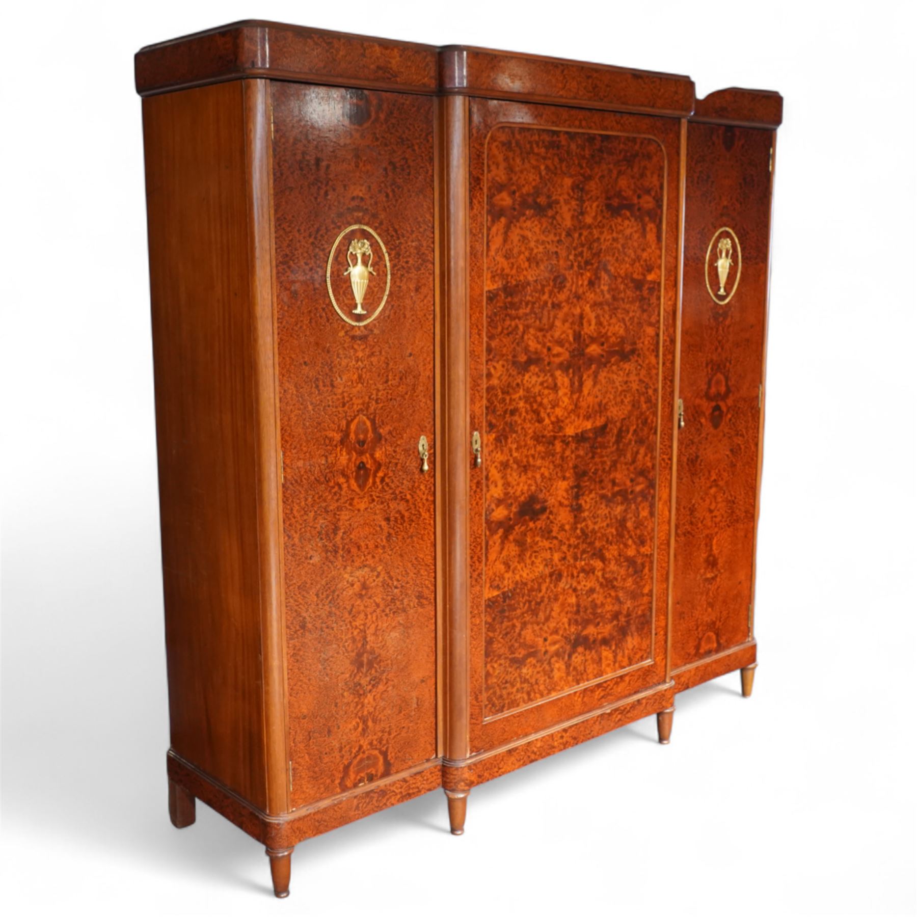 Early 20th century French burr maple breakfront armoire, enclosed by three panelled doors with bookmatched veneer and stringing, outer doors inlaid with gilt urn medallions, fitted interior with hanging spaces flanking a shelved compartment, raised on short tapered legs