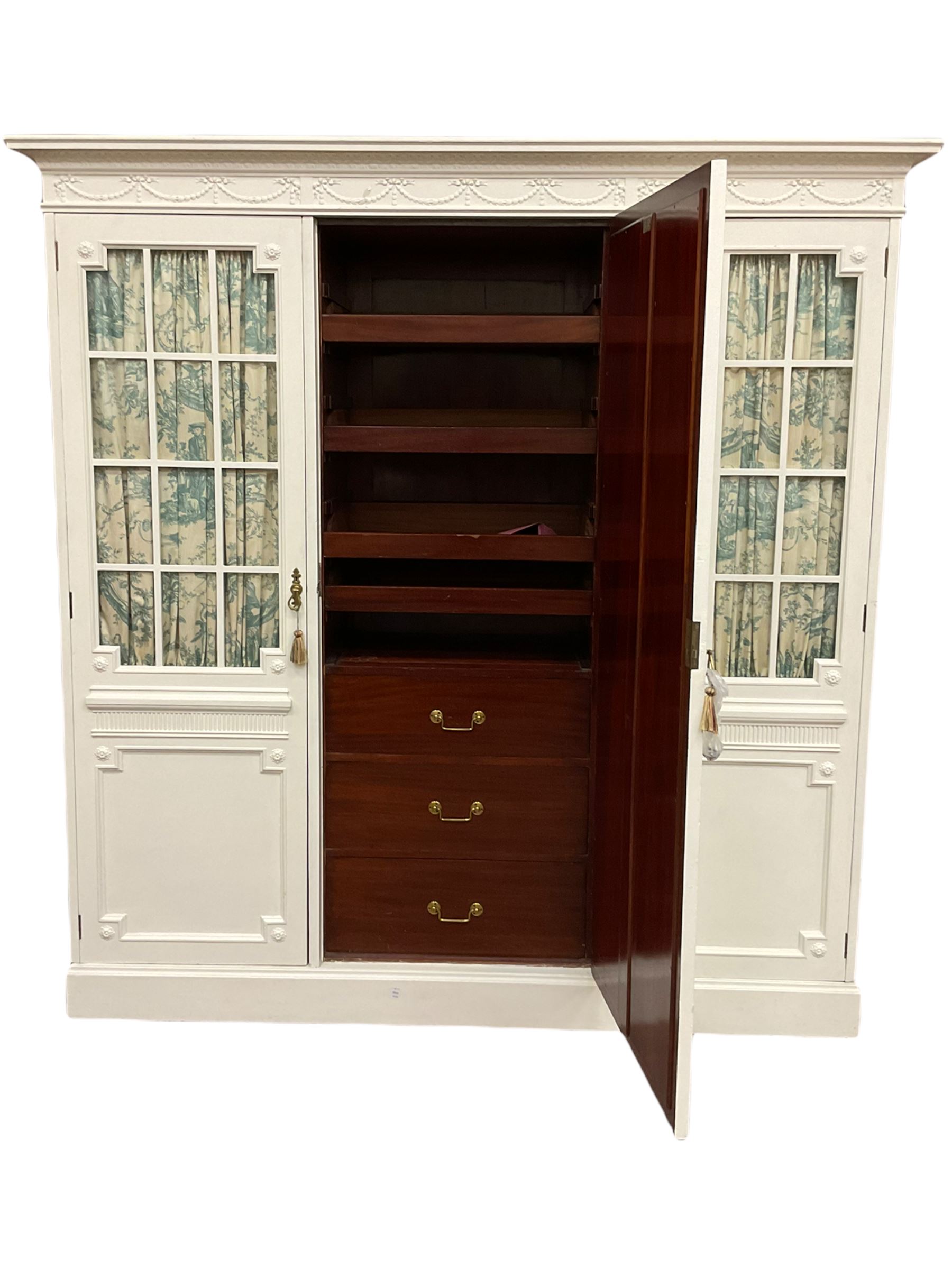 Maple and Co - Edwardian Hepplewhite design white painted triple wardrobe, foliate carved projecting cornice over ribbon and husk swag decorated frieze, two curtain lined astragal glazed doors and centre full mirror door, fitted mahogany interior including slides and drawers, plinth base, label to top of central door