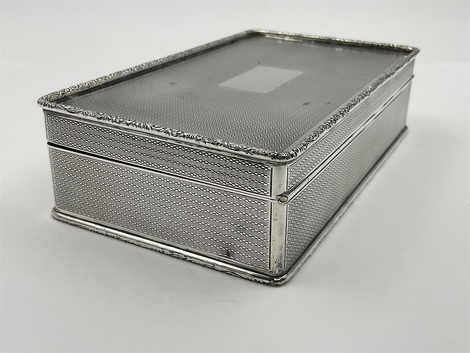 1920's silver table cigarette box, of rectangular form with engine turned decoration to the exterior, the hinged cover with central vacant rectangular panel and raised foliate rim, opening to reveal a gilt interior with later plush lining, hallmarked Charles Henry Dumenil, London 1923, H3.5cm L14.5cm D8.5cm