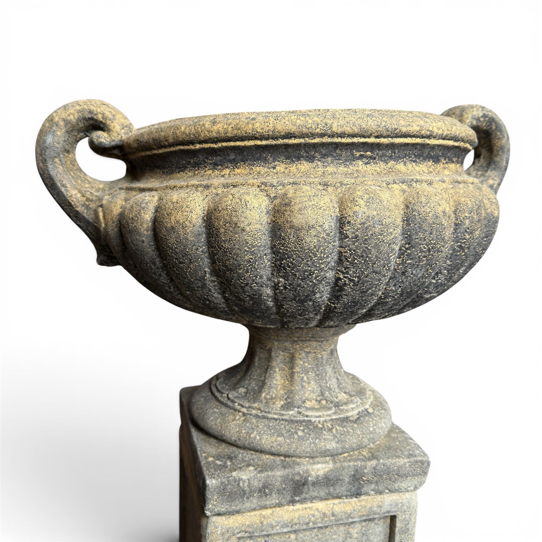 Pair of Neoclassical design weathered cast stone garden urns of kylix form, the rolled rim flanked by twin pierced scroll handles over a shallow gadrooned bowl, raised on a fluted circular socle and faux panelled square pedestal base