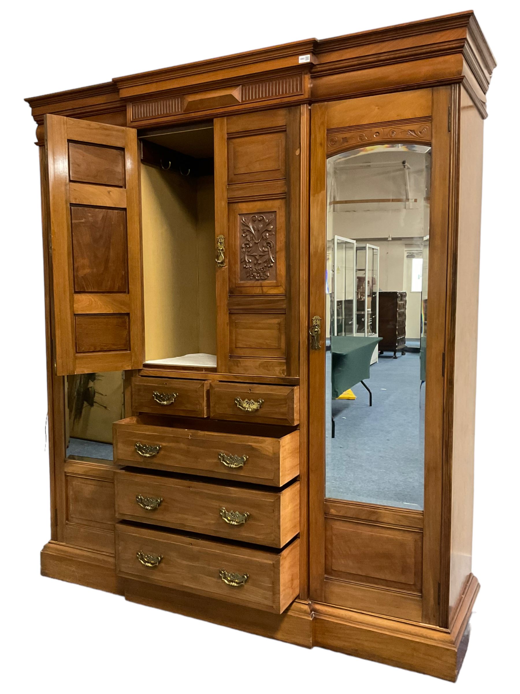Edwardian walnut combination breakfront triple wardrobe, projecting cornice over cushioned and fluted frieze, central fielded panelled cupboard carved with foliate decoration above two short and three long drawers, flanked by two bevelled mirror doors enclosing hanging hooks with single drawers to base, retailed by J Tonks & Sons, Scarborough