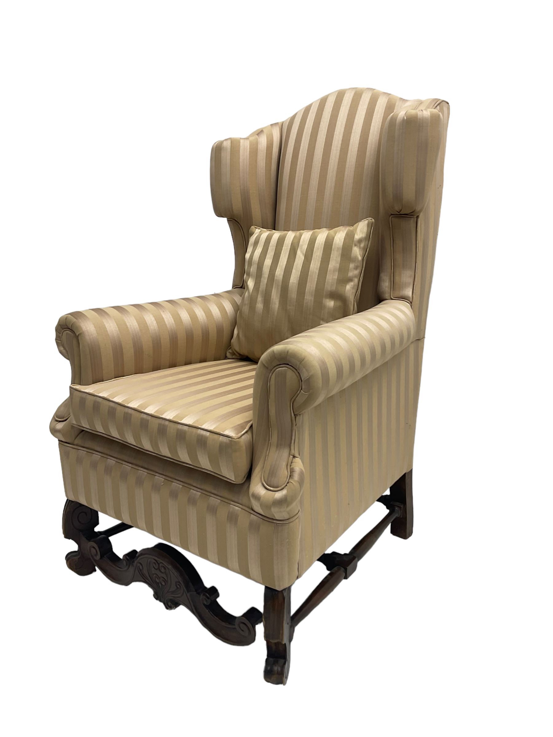 William and Mary style wingback armchair, upholstered in gold striped fabric, on walnut scroll carved supports joined by scroll and foliate carved middle rail, turned H-shaped stretchers, on castors