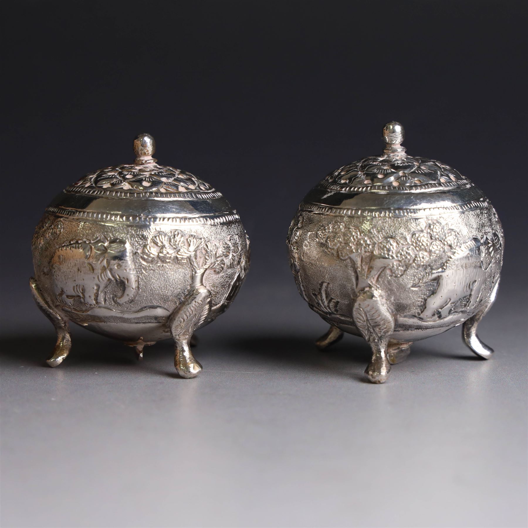 Pair of Middle Eastern silver cruets, of spherical form with pierced tops, embossed with figures, elephants and village scenes, unmarked, together with a pair of silver condiment spoons embossed with floral decoration 
