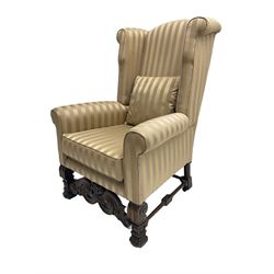 William and Mary style wingback armchair, upholstered in gold striped fabric, on walnut scroll carved supports joined by scroll and foliate carved middle rail, turned H-shaped stretchers, on castors