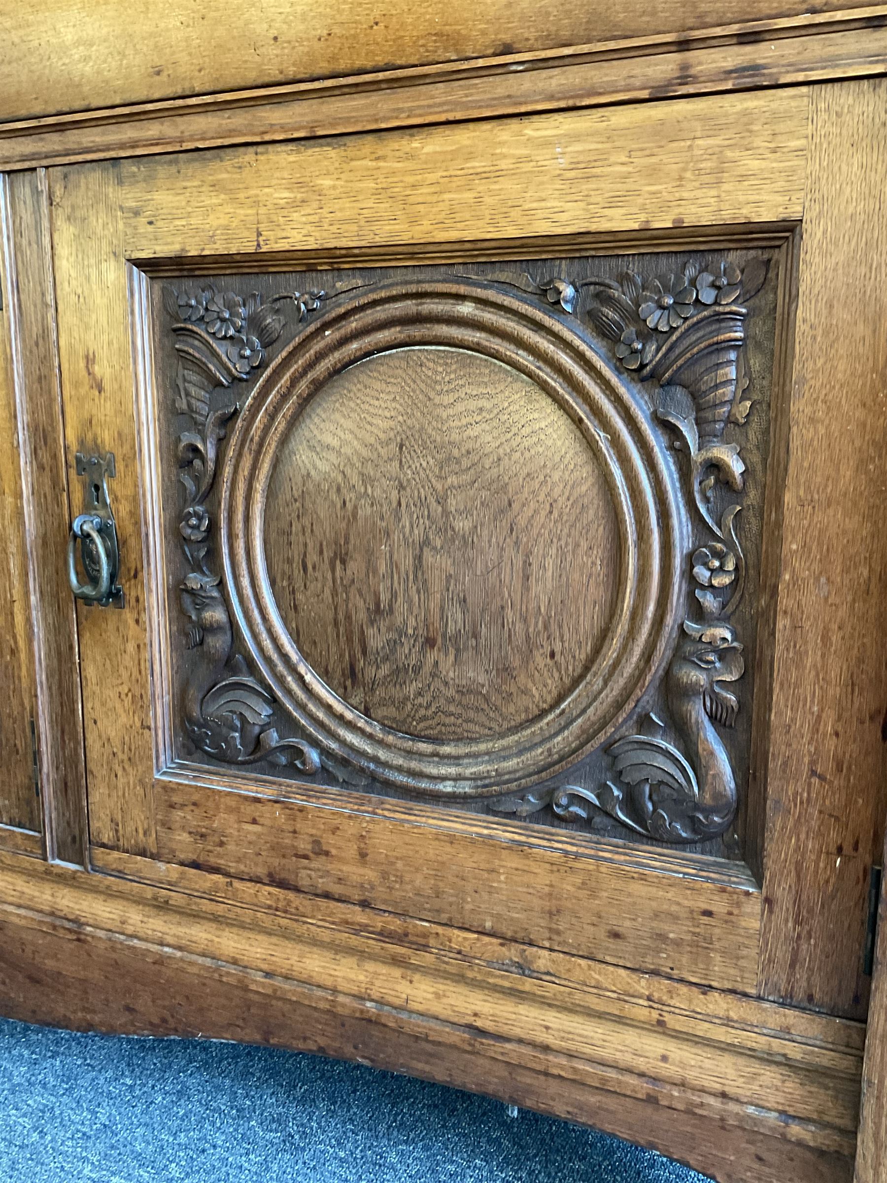 Late 19th century Arts & Crafts period oak mirror back sideboard, shaped bevelled mirror back carved with foliate scrolls and flower heads, down swept supports with elongated club-shaped uprights, moulded rectangular top over two drawers and three cupboards, the drawer fronts with copper handles decorated with sailing ships, panelled doors carved with mythical beasts, flowerheads and foliage, on moulded block feet