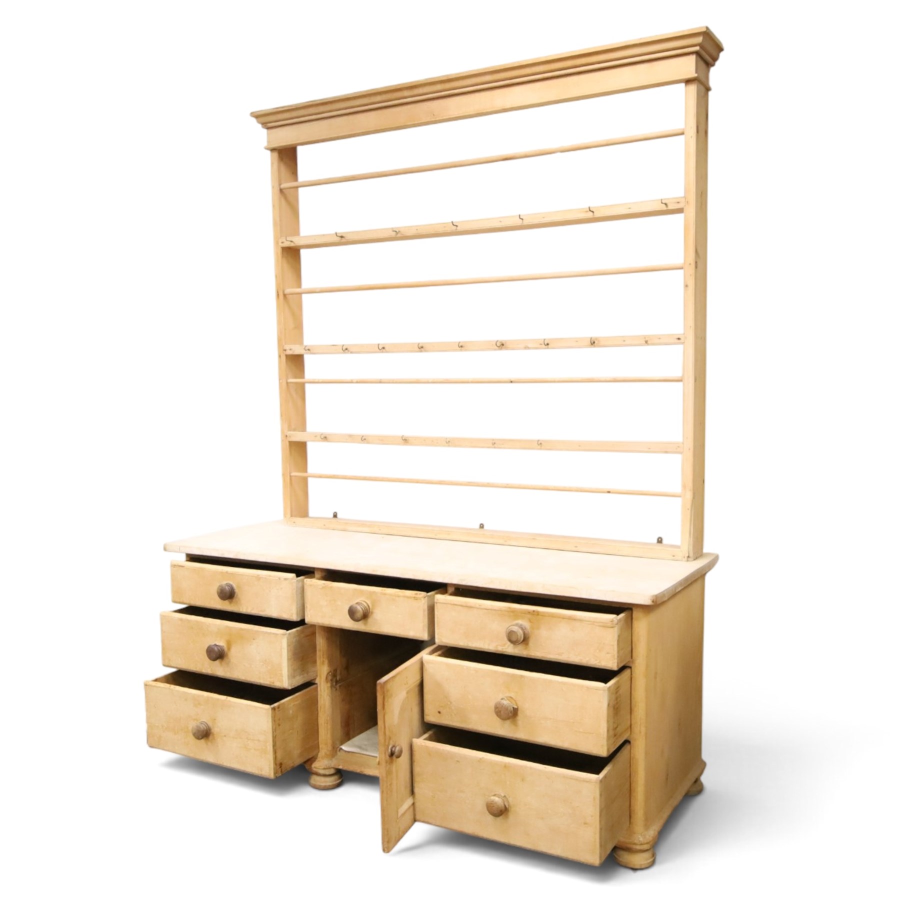 Victorian stripped pine dresser base with plate rack, the four-tier plate rack with supporting rails and hooks, the inverted-breakfront base fitted with seven assorted graduating drawers surrounding a recessed cupboard, on compressed bun feet with castors