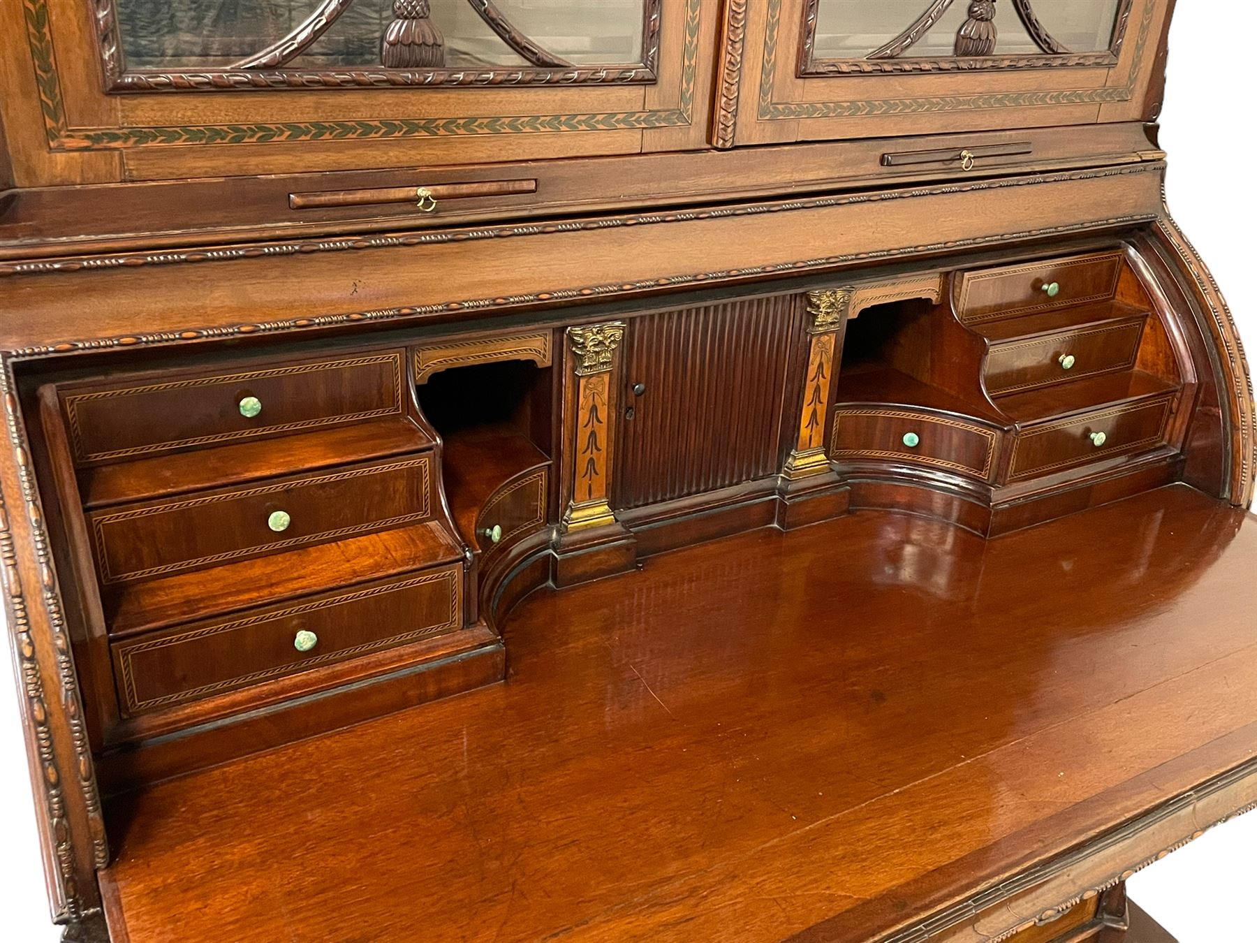Large and impressive late 19th century Irish mahogany secretaire bookcase, moulded sloped arch dentil pediment decorated with central carved mask surrounded by linen festoons and trailing bellflowers, canted and fluted uprights with carved acanthus leaf upper and lower brackets, enclosed by two astragal glazed doors in the form of trophies with twist ribbon carved mouldings, inlaid with fanned spandrels and trailing leaf painted satinwood band, cylinder roll top inlaid with central fan motif and fanned spandrels, pull-out action revealing stepped fitted interior, central tambour roll cupboard flanked by two Composite pilasters and a combination of drawers and pigeon holes, fitted with three break-front drawers, matched canted and fluted corners with acanthus leaf carvings, on square tapering and fluted spade feet