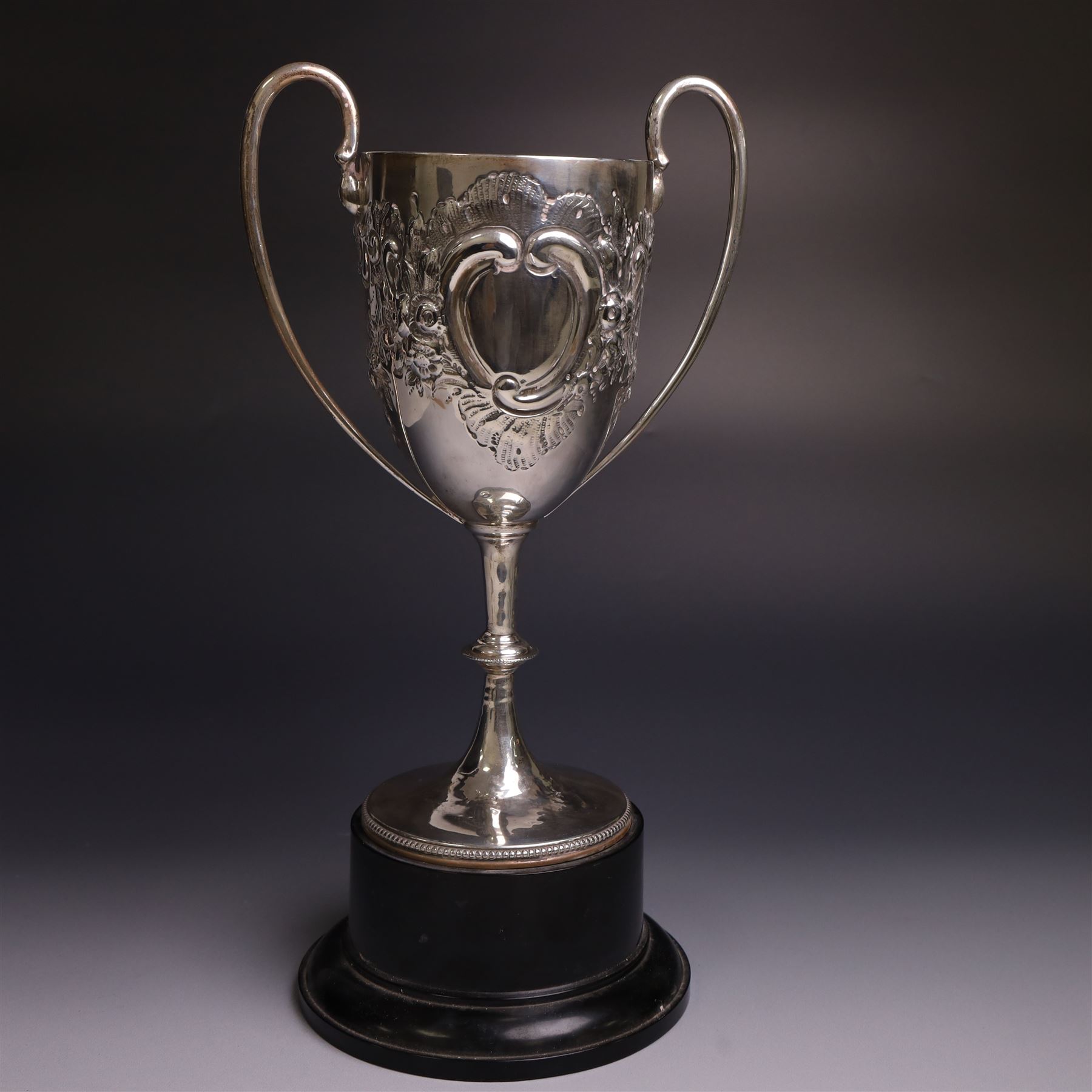 Edwardian silver twin-handled trophy, with embossed floral and scrolling decoration, upon a knopped stem and circular foot, hallmarked Goldsmiths & Silversmiths Co Ltd, London 1903, upon a black plinth, including plinth and handles H32cm