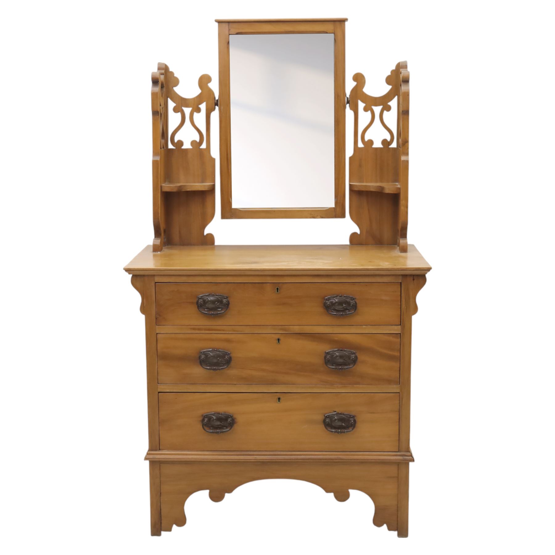 Edwardian stained pine dressing chest, the moulded top over three graduating drawers with pressed brass handles, shaped apron and bracket feet, the mirror back with rectangular plate on pivot supports, flanked by pierced Gothic style uprights with small corner shelves