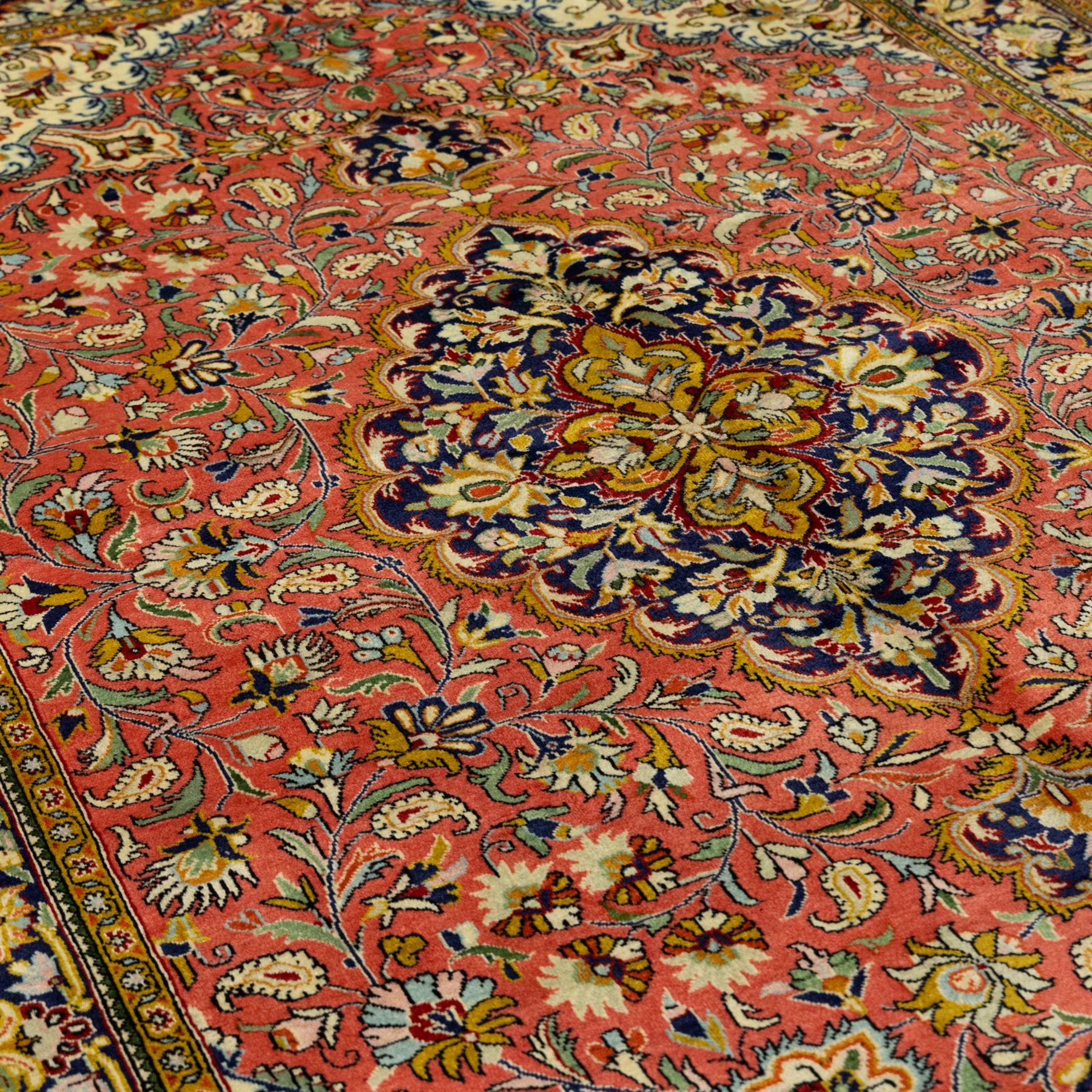 Central Persian Qom hand-knotted carpet, salmon pink ground, with a central dark navy shaped floral medallion with pendants, surrounded by dense scrolling foliate and floral motifs incorporating rosettes, palmettes and delicate vine work, enclosed by ivory spandrels and a dark navy border with repeating palmettes and meandering vines, flanked by narrow floral guard stripes, with cotton fringes