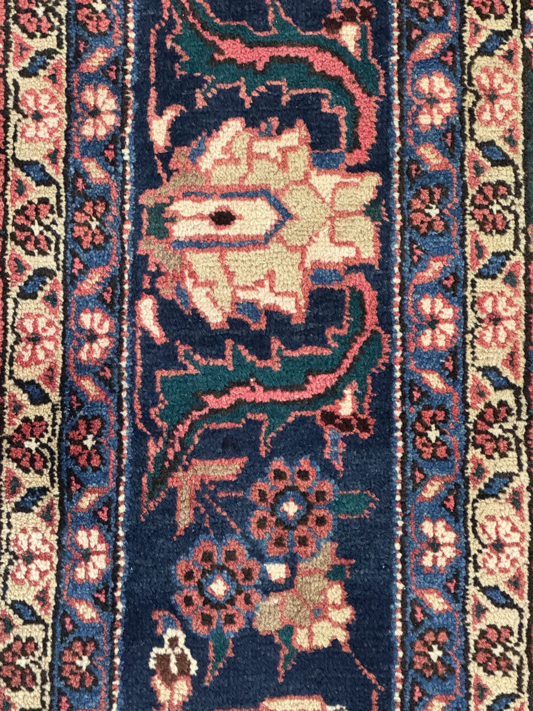 North East Persian Meshed crimson ground carpet, central indigo and green medallion with floral decoration, busy field with trailing foliage and stylised flowers, matching spandrels with floral motifs, indigo border featuring scrolling vines and palmettes, framed by narrow floral guard bands
