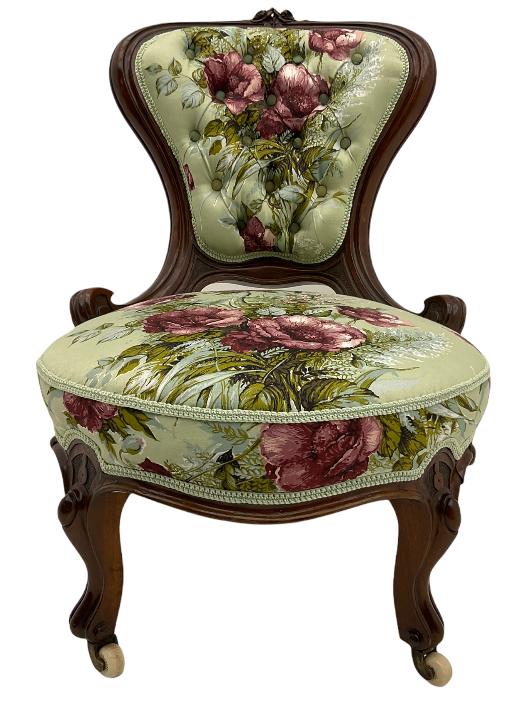 Victorian walnut framed nursing chair, spade shaped back carved with flower heads, upholstered in buttoned rose patterned fabric, on scroll carved cabriole supports terminating at brass and ceramic castors