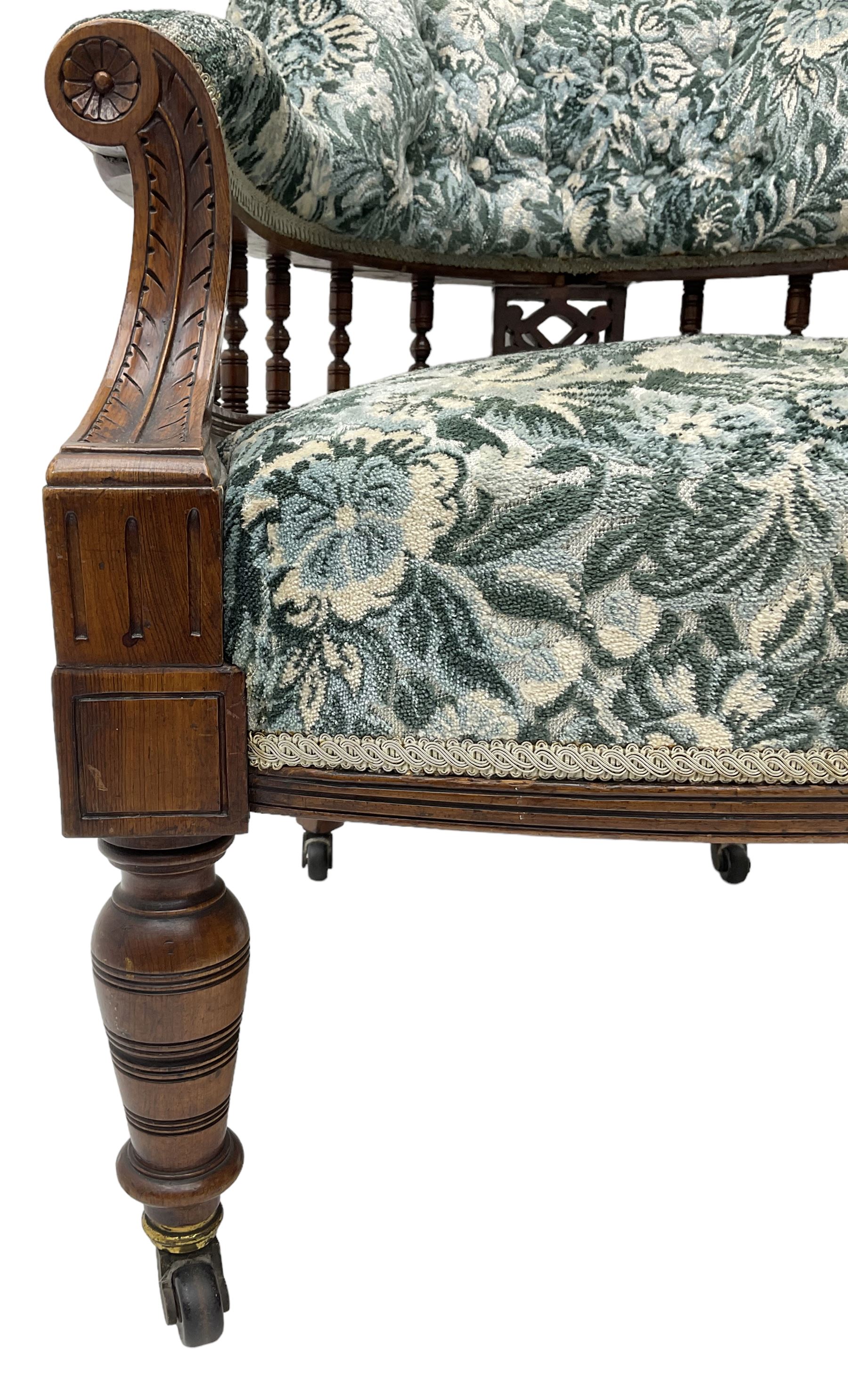Victorian walnut framed tub-shaped armchair, the shaped rolled back supported by a spindle turned balustrade, upholstered in buttoned floral fabric, scrolled arms supports carved with acanthus leaves and flower heads, on collar turned feet with brass and ceramic castors