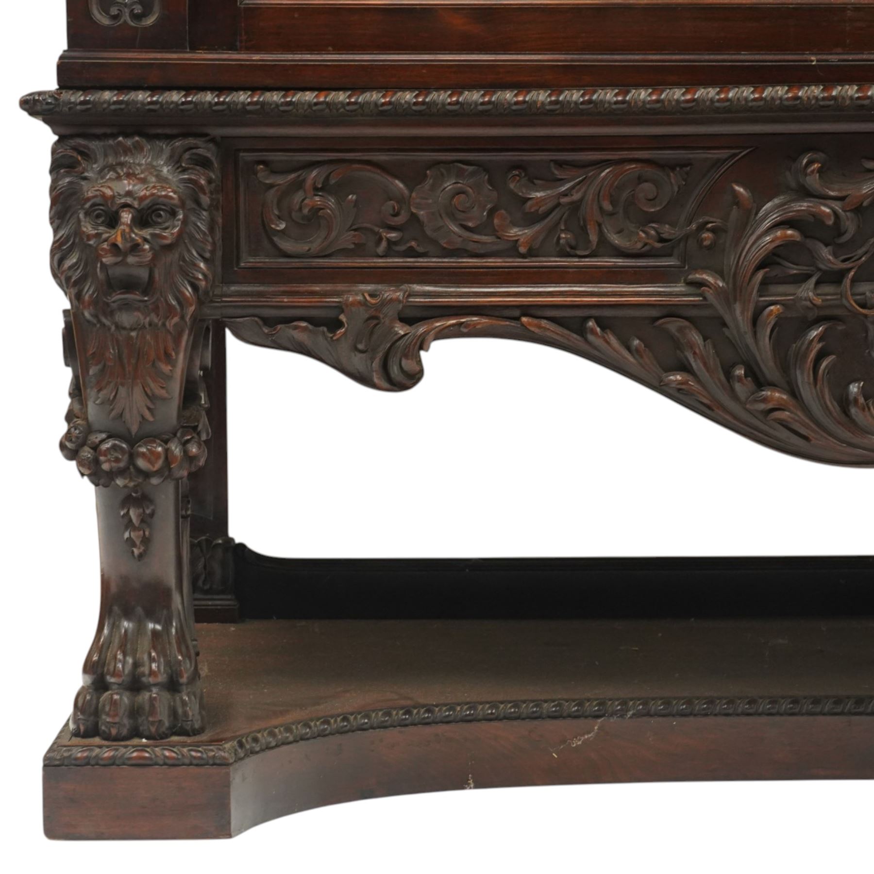 Large and impressive 19th century heavily carved mahogany display cabinet, the ornate pediment carved with a central scalloped and foliate rocaille motif with extending acanthus leaves and egg-and-dart moulding flanked by acanthi finials, over a central cartouche with carved bow-and-arrow motif and trailing scrollwork, the bookcase enclosed by two glazed doors with bevelled glass sides, the base with a gadrooned upper edge over a central rocaille ornament with scrolling acanthus leaves, the panelled frieze with further foliate motifs, raised on large carved snarling lion mask supports with floral garlands and claw feet, united by a concave rectangular platform base with gadrooning egg-and-dart moulding