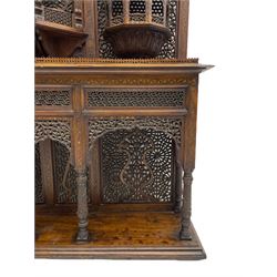Late 19th century Indian Burmese carved and pierced hardwood and brass inlaid wall shelf, the shaped back profusely decorated with trailing and interlaced foliage, with projecting architectural structures, the central balcony with canopy top and five open windows with arched apertures on columns, two flanking five-sided balconies, the shelf supported by six foliate carved columns with pierced cusped pointed arches 