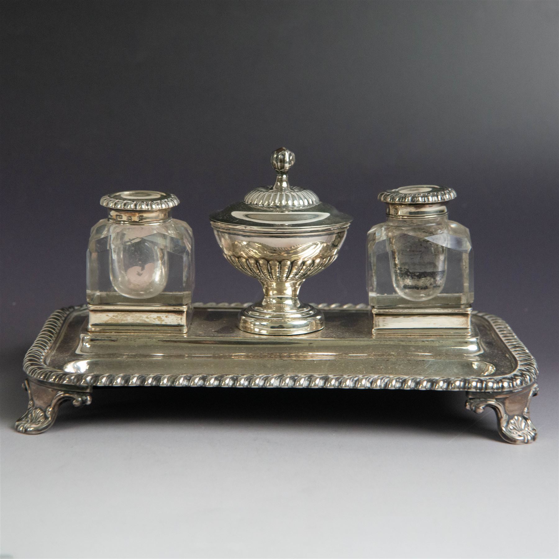 George IV silver desk stand, of rectangular form with rounded corners and oblique gadrooned rim, upon four embossed pad feet, with central pedestal dish of part fluted form with domed lid engraved with crest and with bud finial, with two recesses either side housing faceted glass inkwells, each with silver collars, hallmarked John & Thomas Settle, Sheffield 1825 & 1827, H11cm