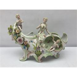 Continental porcelain figural centrepiece, decorated with a female figure and two putti playing instruments, encrusted throughout with flower heads, together with a similar jar and cover with peony finial, a chocolate cup with cover and saucer and a quatrefoil cup and saucer, each decorated with figural panels with gilt highlights, both cups in the style of Helena Wolfsohn, centrepiece H21cm
