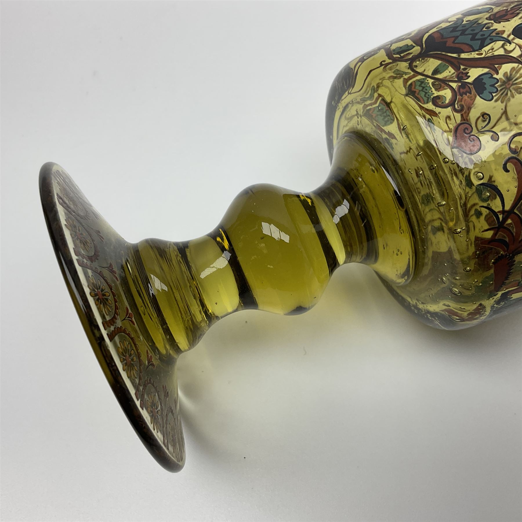 19th Century Bohemian amethyst glass lidded goblet, the bowl of faceted form, gilded with three children in period dress playing games, upon a knopped stem and conforming faceted foot, together with an art nouveau green glass goblet, with gilded rim, upon clear trumpet stem, and a hand blown wine glass, transfer printed with birds and three figures in period dress, tallest H29cm