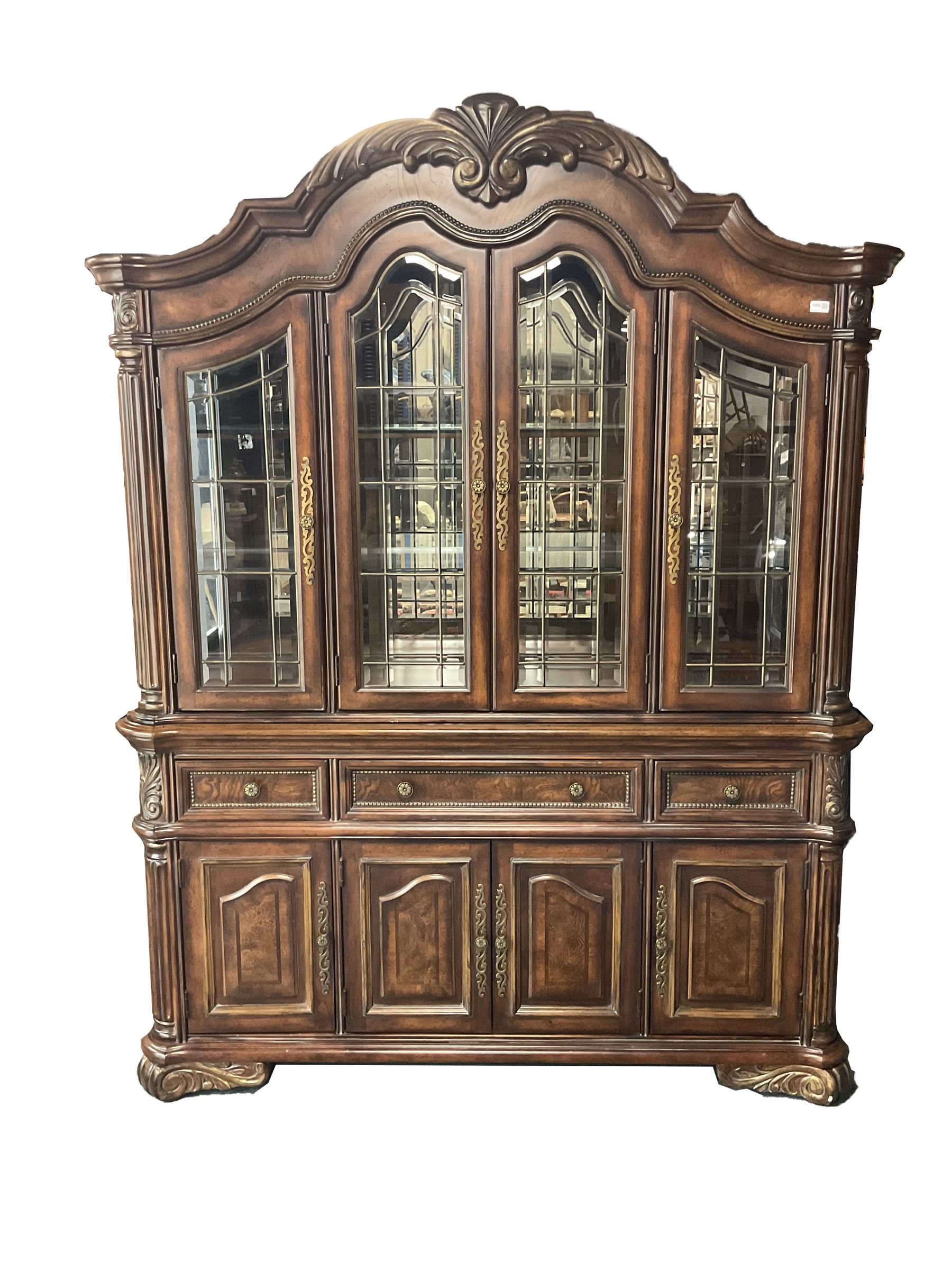Kevin Charles American walnut display cabinet, illuminated interior, shell carved pediment above four bevel edge doors enclosing six glazed shelves above one central long drawer flanked by two short drawers, above three cupboards, carved bracket supports 