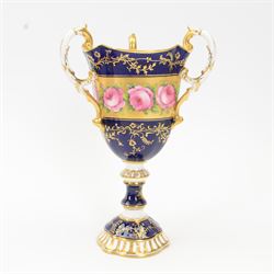 Early 19th century Coalport three handled goblet vase, shape no 114, and pair of plates, each hand painted with flowering pink roses upon a gilt band, against a dark blue ground heightened with trailing gilt tendrils, each marks beneath Coalport Swansea Period 1818, including handles vase H22.5cm, plates D23cm