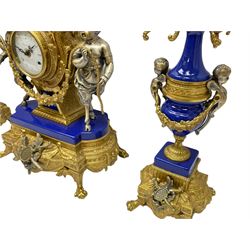 A 20th century continental gilt metal Lyre mantle clock with a pair of matching six light candelabra, spring driven movement housed in a gilt drum case on a brass and cobalt blue base with paw feet, two bronzed metal figures of mythological cherubs with animal legs and cloven hooves supporting a festooned garland swag, eight-day twin barrel striking movement with a floating lever balance escapement, striking the hours and half-hours on two bells, white enamel dial with roman numerals, minute track and pierced steel hands, dial inscribed “Imperial”. 