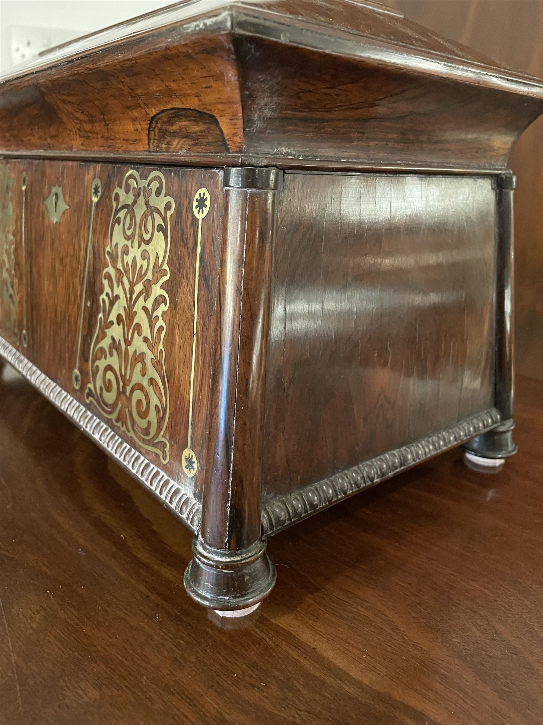 Early 19th century rosewood and brass inlaid tea caddy, of sarcophagus form, the front and hinged opening cover inlaid with brass scrolling panels, opening to reveal a compartmented interior with twin removable boxes with conforming inlaid brass detail to the hinged covers and zinc lining to the interiors, flanking a later glass mixing bowl, the whole upon four turned cylindrical feet, H21cm W36cm D20cm