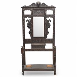 Late Victorian heavily carved oak hall stand, the scrollwork pediment with carved lion's head, bevelled mirror plate flanked by carved dragon motifs, over hinged storage compartment and umbrella stands with drip trays, on turned front supports