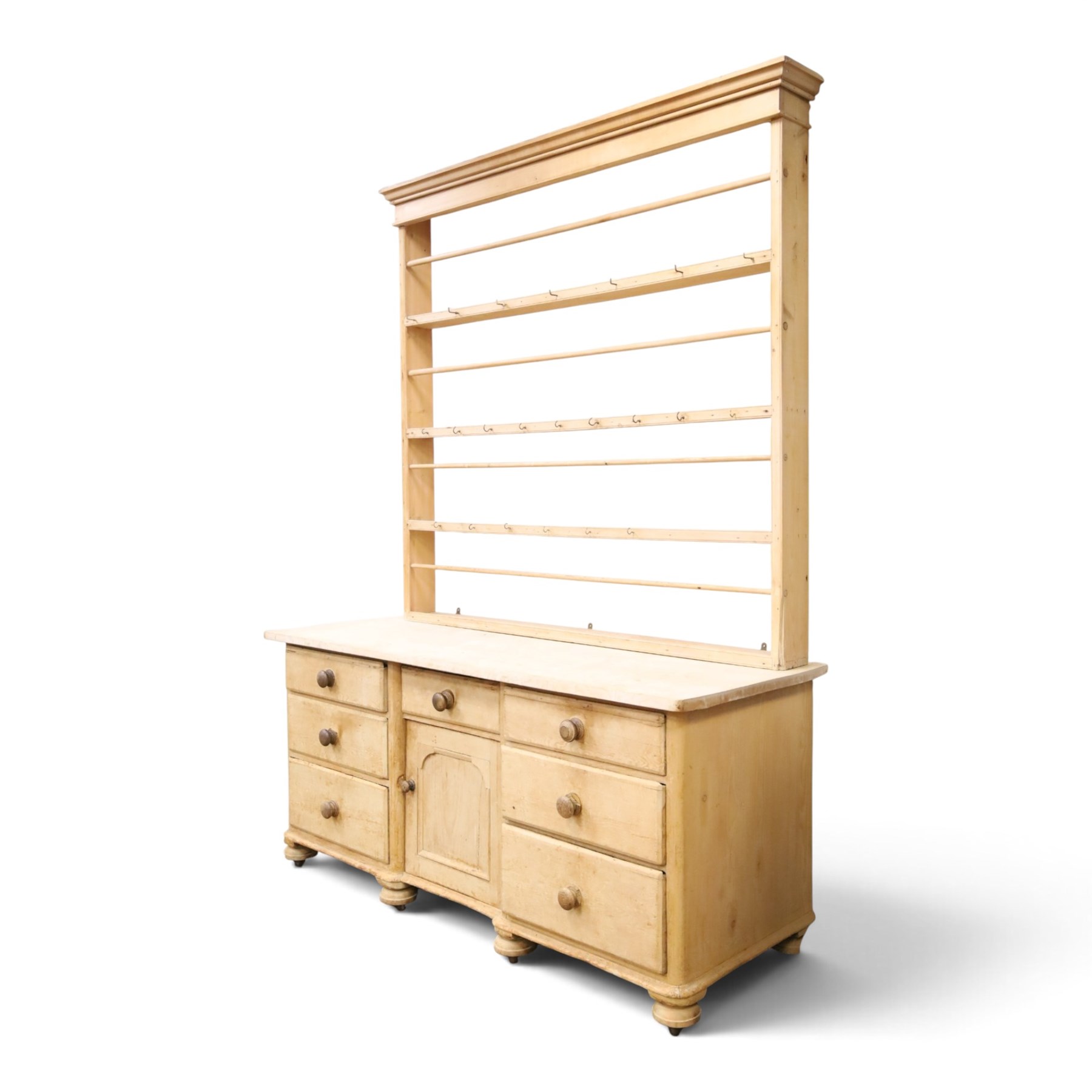 Victorian stripped pine dresser base with plate rack, the four-tier plate rack with supporting rails and hooks, the inverted-breakfront base fitted with seven assorted graduating drawers surrounding a recessed cupboard, on compressed bun feet with castors