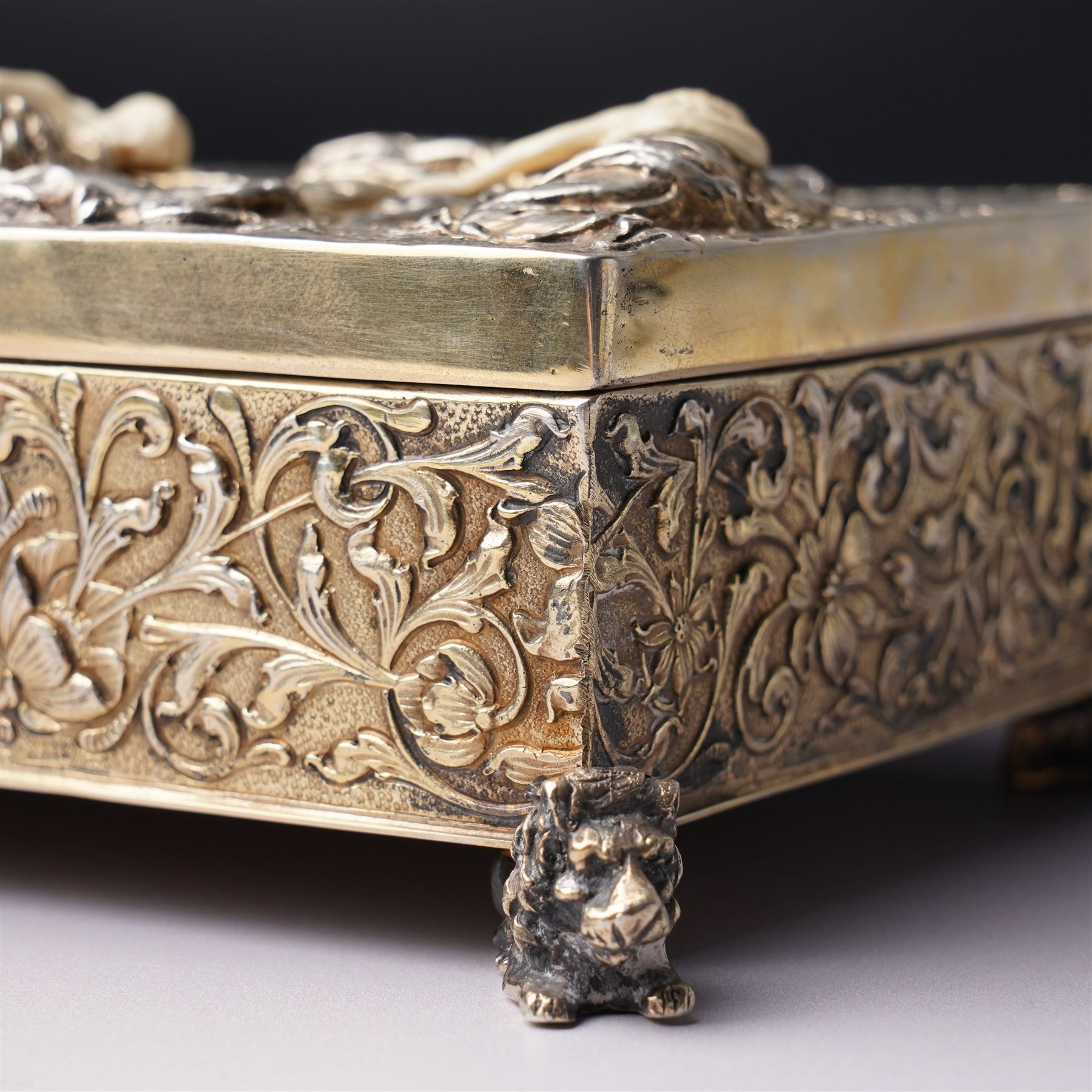 German 800 standard silver gilt and ivory mounted box, the hinged cover embossed and chased with an allegorical feasting scene, the sides with floral decoration, on four cast lion feet 21cm x 16cm x 8cm Maker Ludwig Neresheimer, Hanau circa 1900 This item has been registered for sale under Section 10 of the APHA Ivory Act