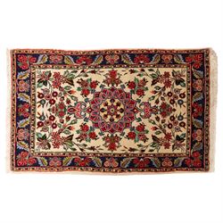 Two small rugs, an ivory ground example decorated with a central floral medallion and scattered rose sprays, enclosed by a navy foliate border (62cm x 101cm), and a crimson pictorial mat worked with seated figures, an animal and birds amongst flowering plants (45cm x 79cm) (2)