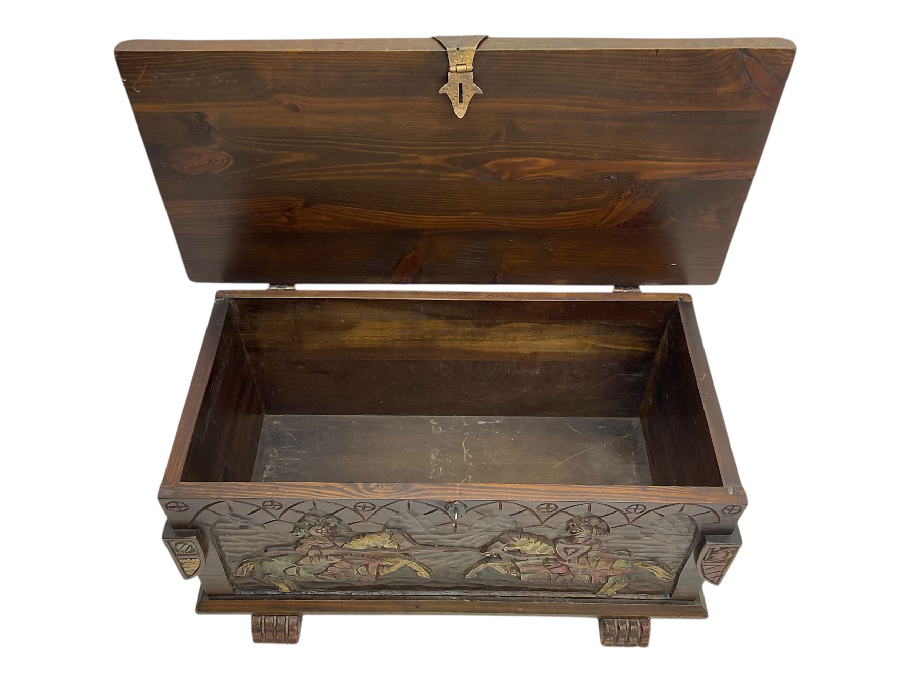 Mid-20th century carved hardwood chest, the rectangular hinged top with decorative strap hinges, the front and sides deeply carved with medieval-style jousting knights, heraldic shields, and architectural elements, raised on shaped block feet