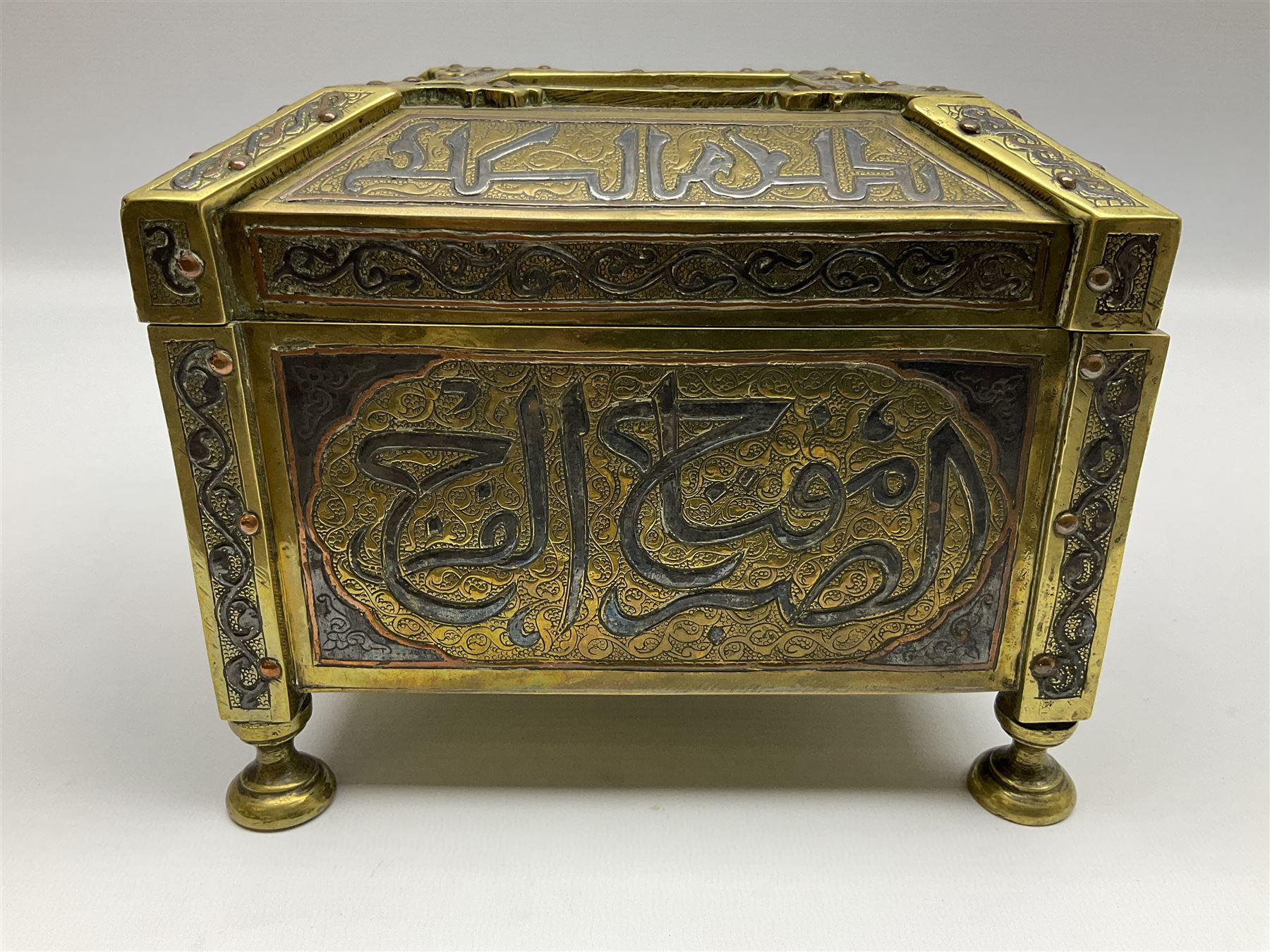 Late 19th century Damascus Cairoware Quran box, of square form with canted hinged cover, upon four circular pad feet, the brass exterior inlaid with silver and copper script and decorative detail, opening to reveal a marquetry lined interior, H15.5cm W19cm D19cm