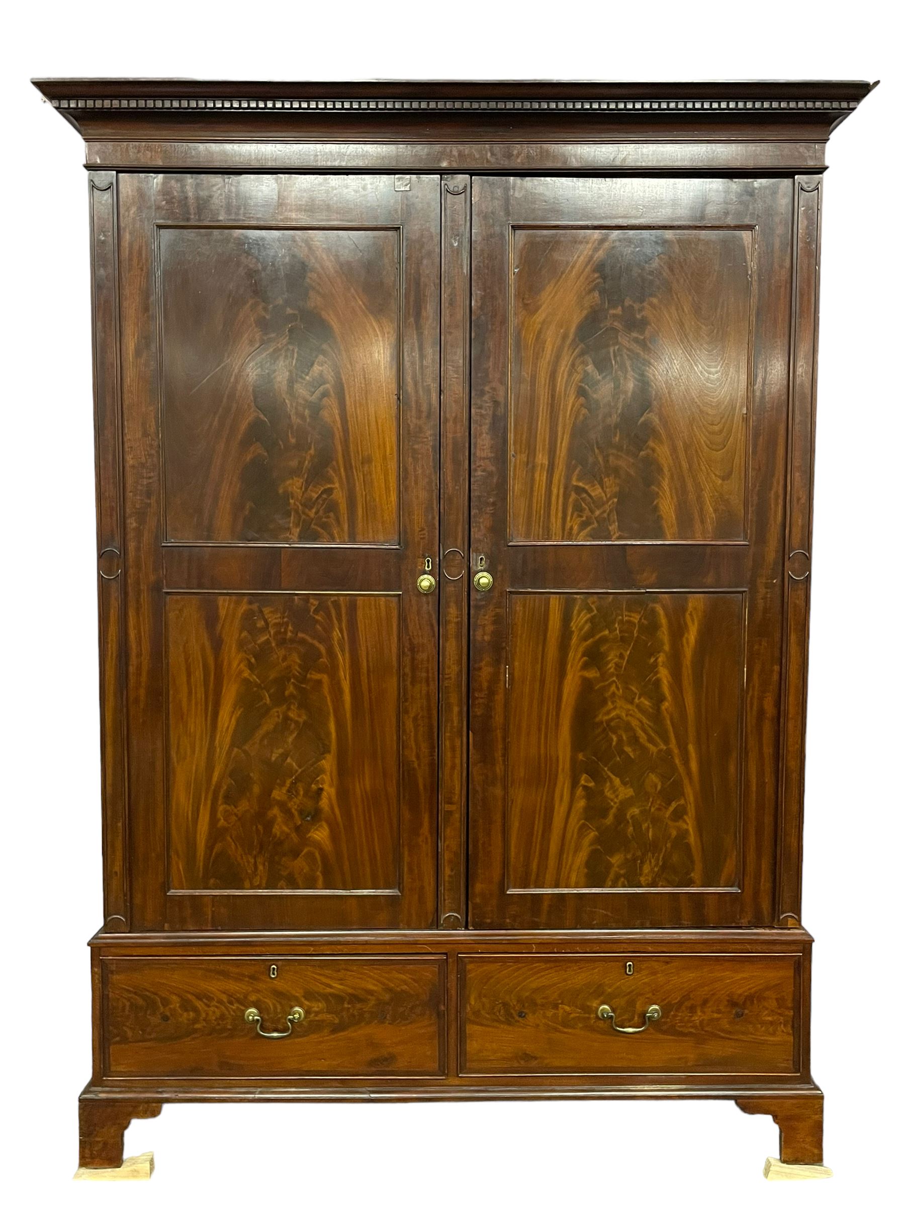 George III mahogany double wardrobe, the projecting cornice with dentil moulding over two panelled doors with mirror-lined interiors, enclosing hanging rails and brass hooks, the base fitted with two drawers, raised on bracket feet